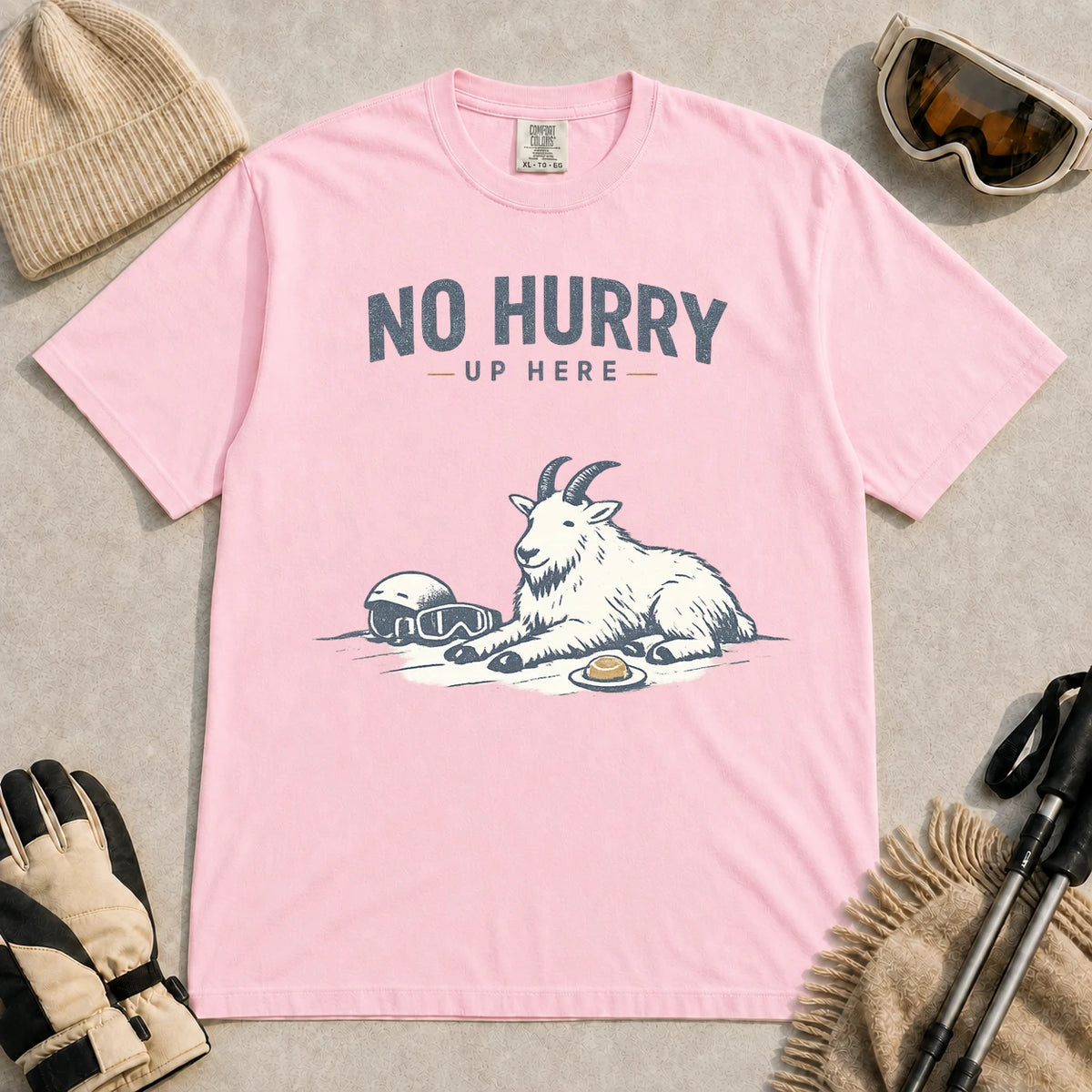 Blossom tee featuring a relaxed mountain goat resting in the snow beside ski goggles and a helmet with the phrase “No Hurry Up Here.”