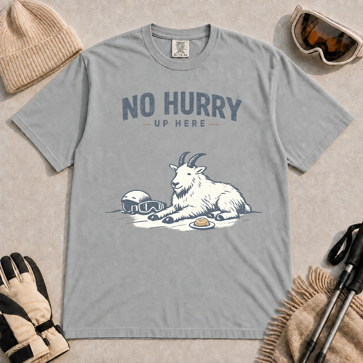 Granite tee featuring a relaxed mountain goat resting in the snow beside ski goggles and a helmet with the phrase “No Hurry Up Here.”