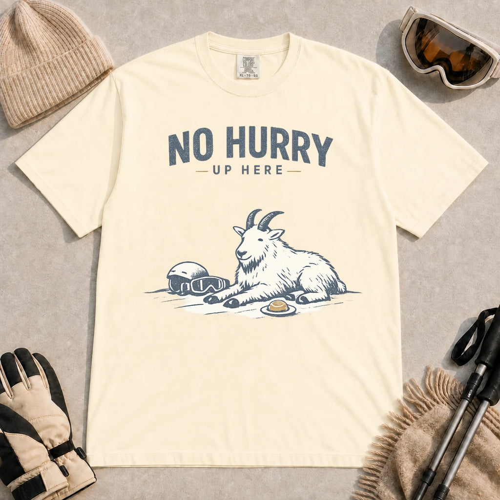 Ivory tee featuring a relaxed mountain goat resting in the snow beside ski goggles and a helmet with the phrase “No Hurry Up Here.”