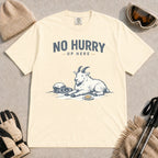 Ivory tee featuring a relaxed mountain goat resting in the snow beside ski goggles and a helmet with the phrase “No Hurry Up Here.”