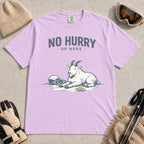 Orchid tee featuring a relaxed mountain goat resting in the snow beside ski goggles and a helmet with the phrase “No Hurry Up Here.”