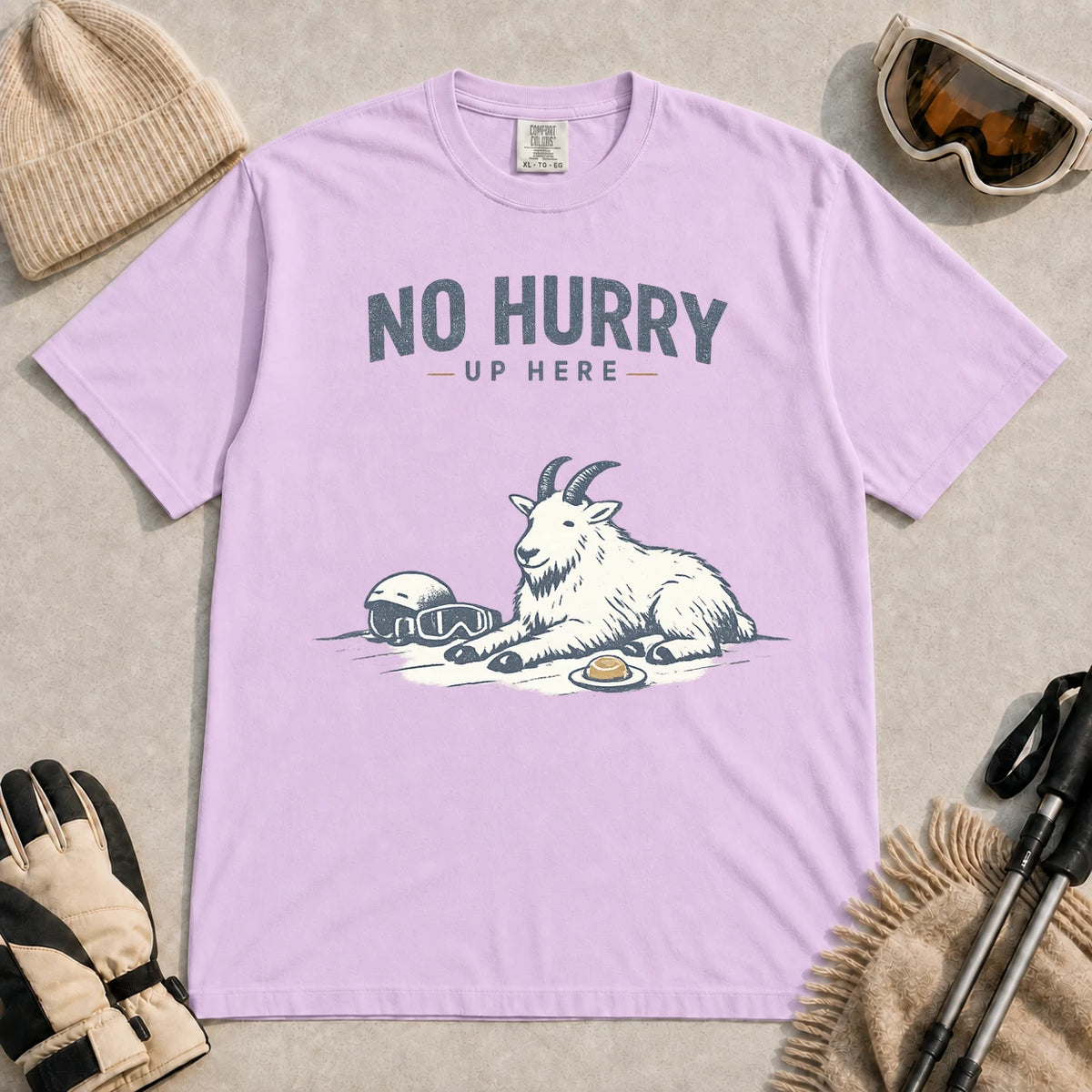 Orchid tee featuring a relaxed mountain goat resting in the snow beside ski goggles and a helmet with the phrase “No Hurry Up Here.”