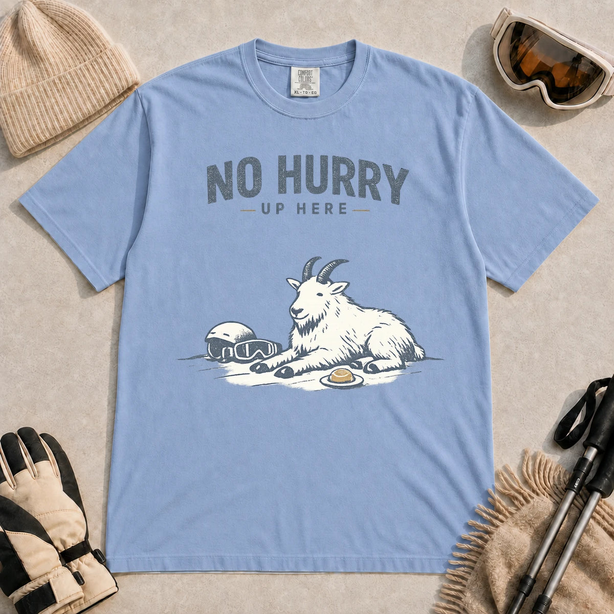 Washed Denim tee featuring a relaxed mountain goat resting in the snow beside ski goggles and a helmet with the phrase “No Hurry Up Here.”