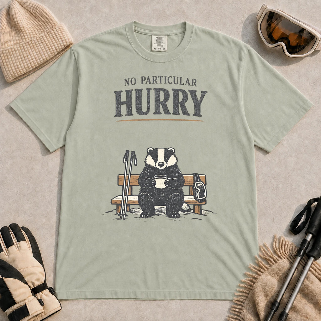 Bay tee featuring a relaxed badger sitting on a snowy bench with a mug, ski poles, and goggles, with the phrase “No Particular Hurry.”