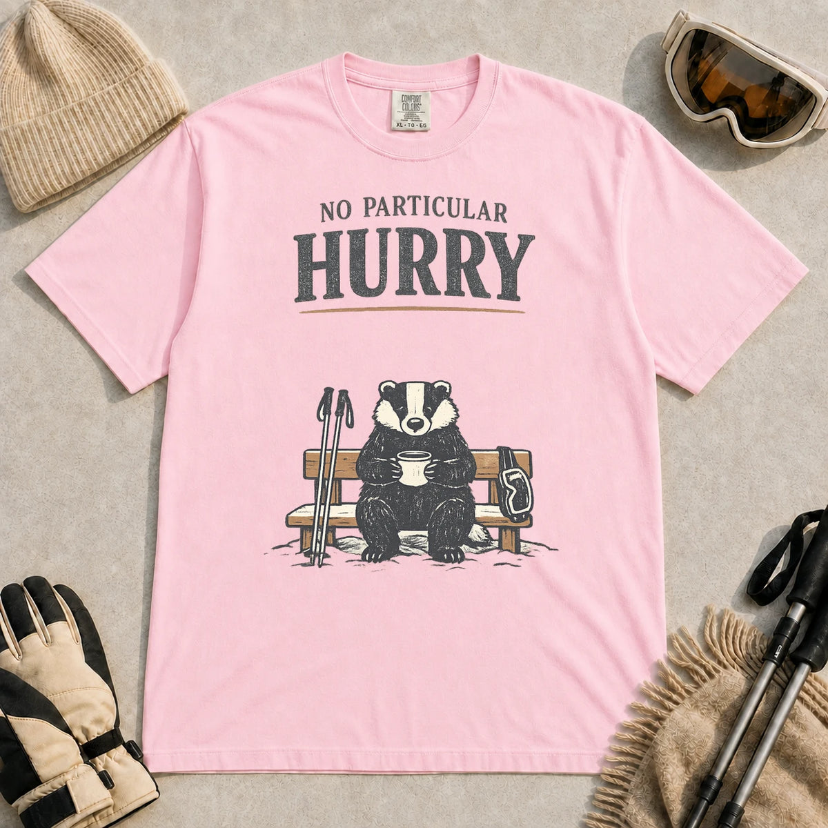 Blossom tee featuring a relaxed badger sitting on a snowy bench with a mug, ski poles, and goggles, with the phrase “No Particular Hurry.”