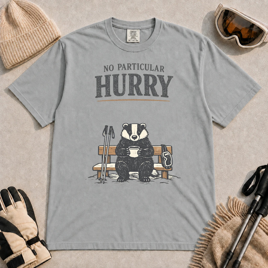 Granite tee featuring a relaxed badger sitting on a snowy bench with a mug, ski poles, and goggles, with the phrase “No Particular Hurry.”
