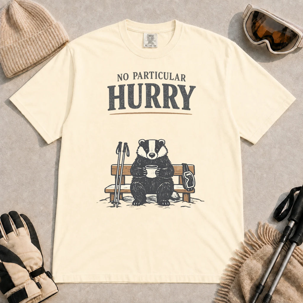 Ivory tee featuring a relaxed badger sitting on a snowy bench with a mug, ski poles, and goggles, with the phrase “No Particular Hurry.”
