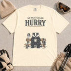 Ivory tee featuring a relaxed badger sitting on a snowy bench with a mug, ski poles, and goggles, with the phrase “No Particular Hurry.”
