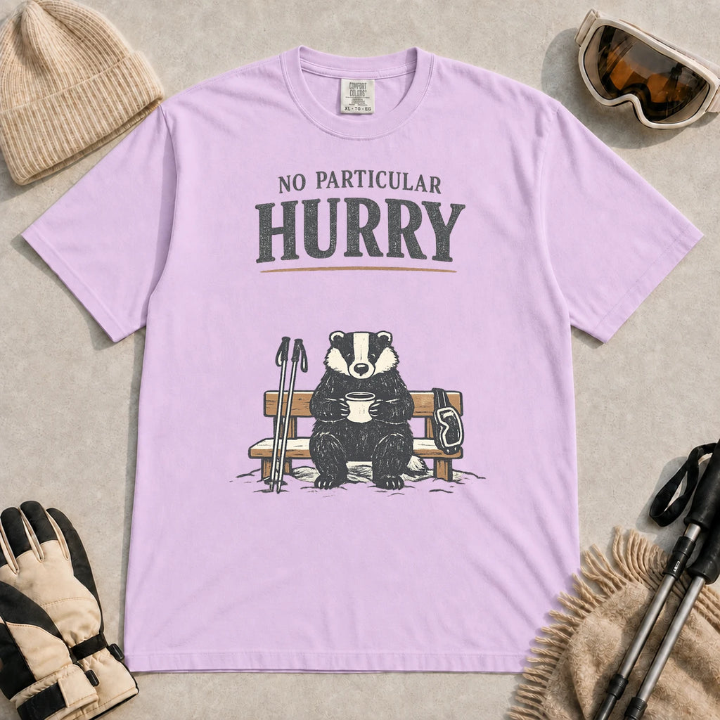Orchid tee featuring a relaxed badger sitting on a snowy bench with a mug, ski poles, and goggles, with the phrase “No Particular Hurry.”
