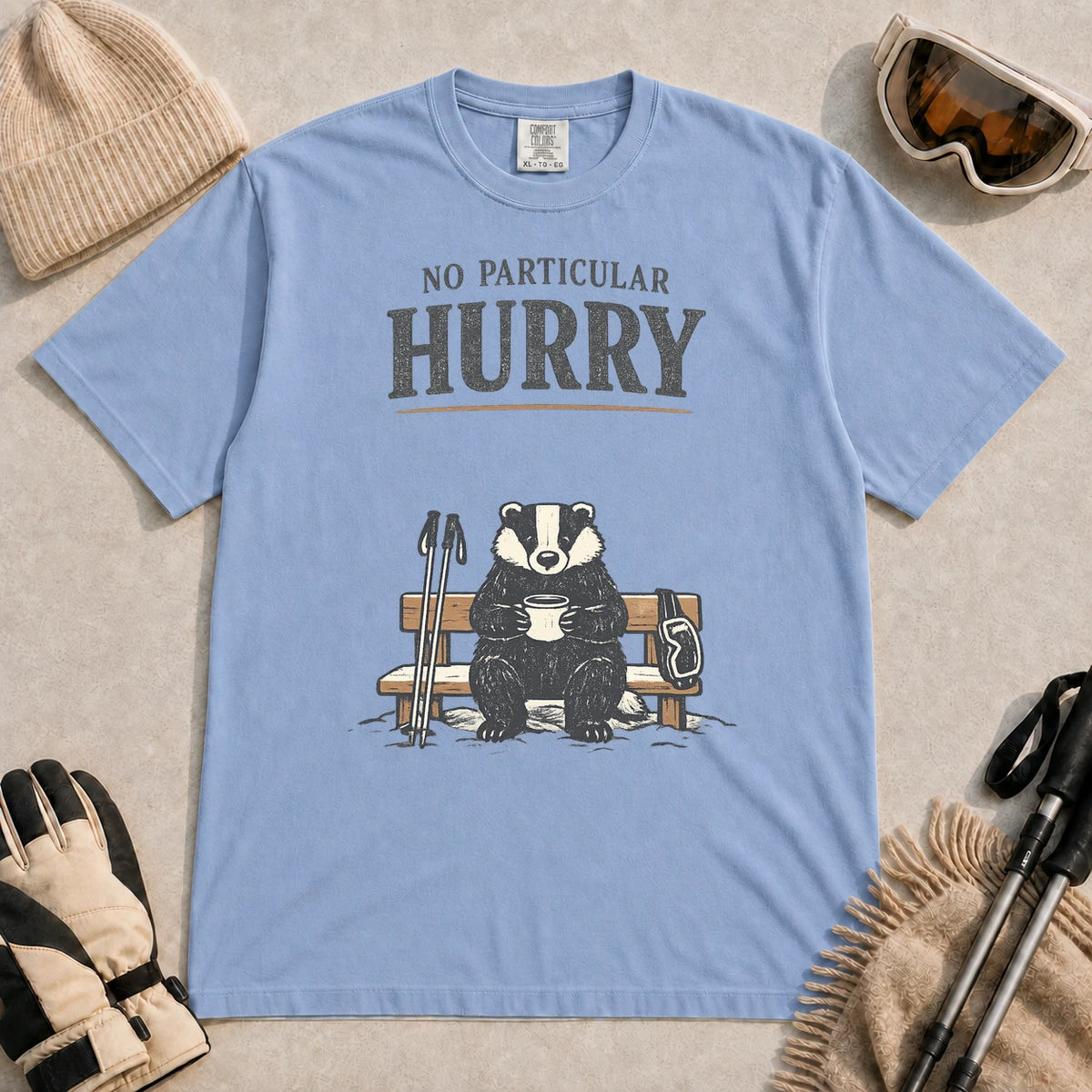 Washed Denim tee featuring a relaxed badger sitting on a snowy bench with a mug, ski poles, and goggles, with the phrase “No Particular Hurry.”