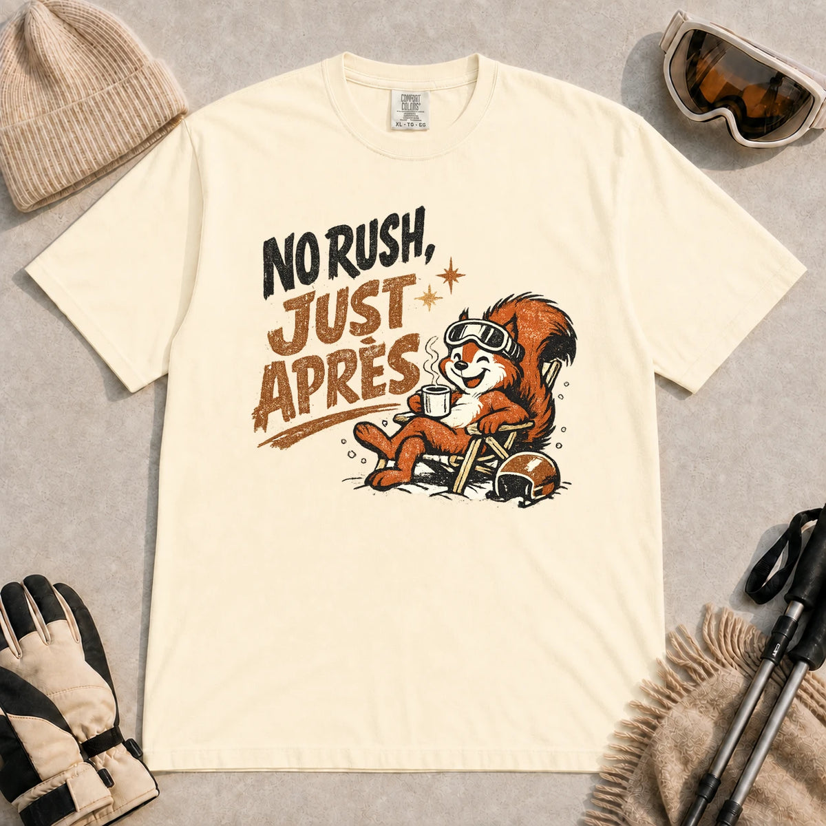 Ivory tee featuring a cheerful squirrel in ski goggles relaxing in a chair with a hot drink and helmet, with the phrase “No Rush, Just Après.”