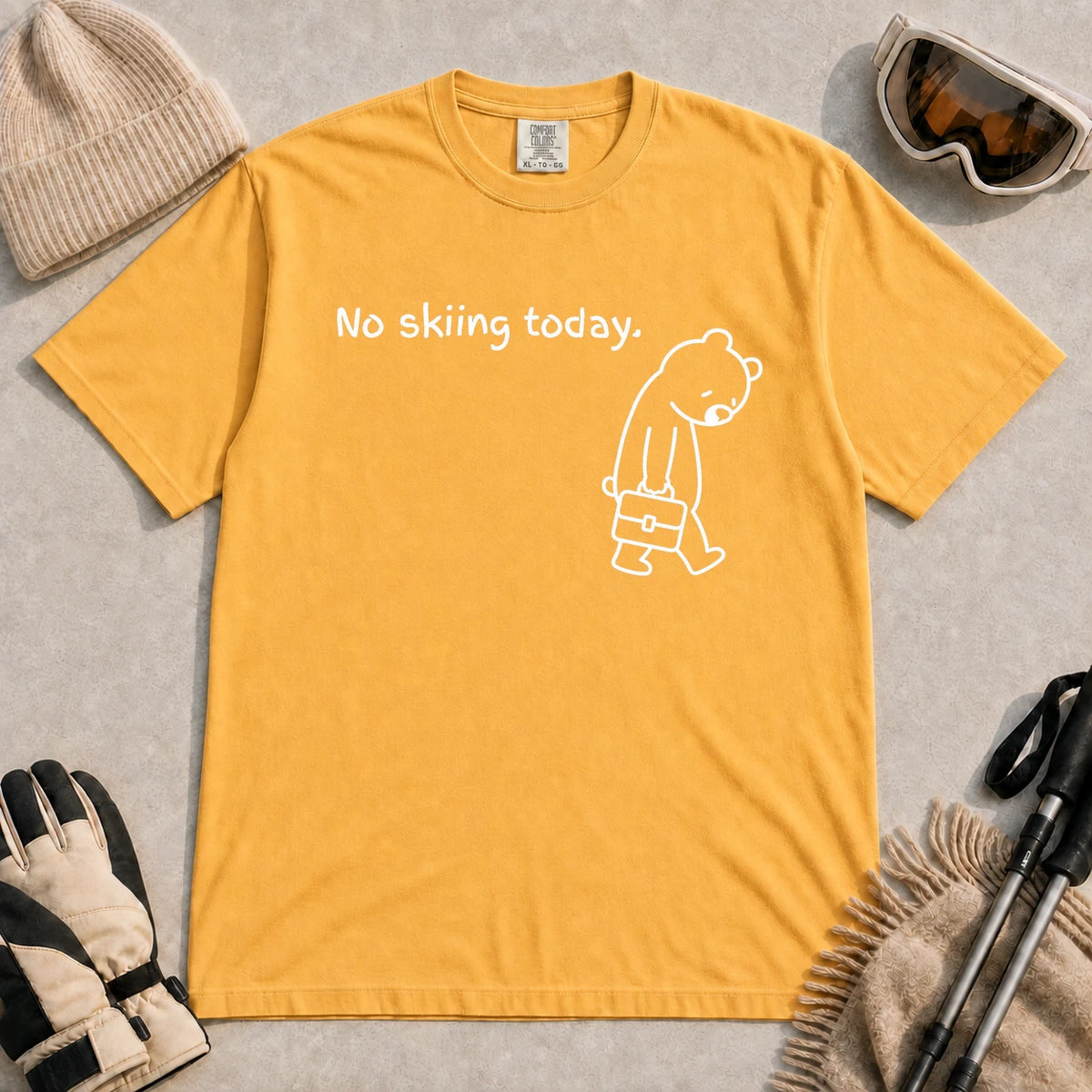 Mustard tee featuring a sad bear dressed for work and carrying a briefcase beneath the phrase “No skiing today.”