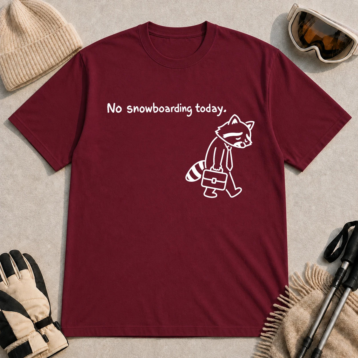 Maroon tee featuring a sad raccoon dressed for work and carrying a briefcase beneath the phrase “No snowboarding today.”