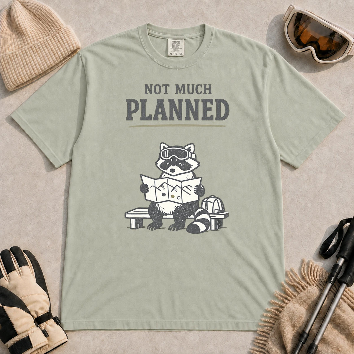 Bay tee featuring a raccoon in ski goggles sitting on a bench and studying a trail map beside a backpack, with the phrase “Not Much Planned.”
