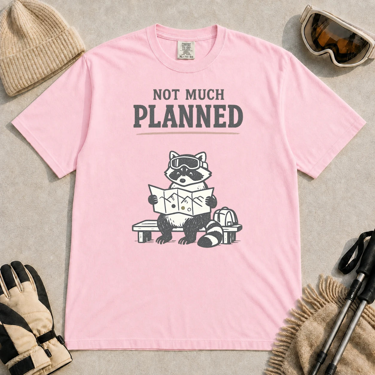 Blossom tee featuring a raccoon in ski goggles sitting on a bench and studying a trail map beside a backpack, with the phrase “Not Much Planned.”