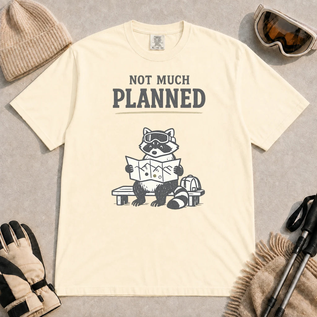 Ivory tee featuring a raccoon in ski goggles sitting on a bench and studying a trail map beside a backpack, with the phrase “Not Much Planned.”