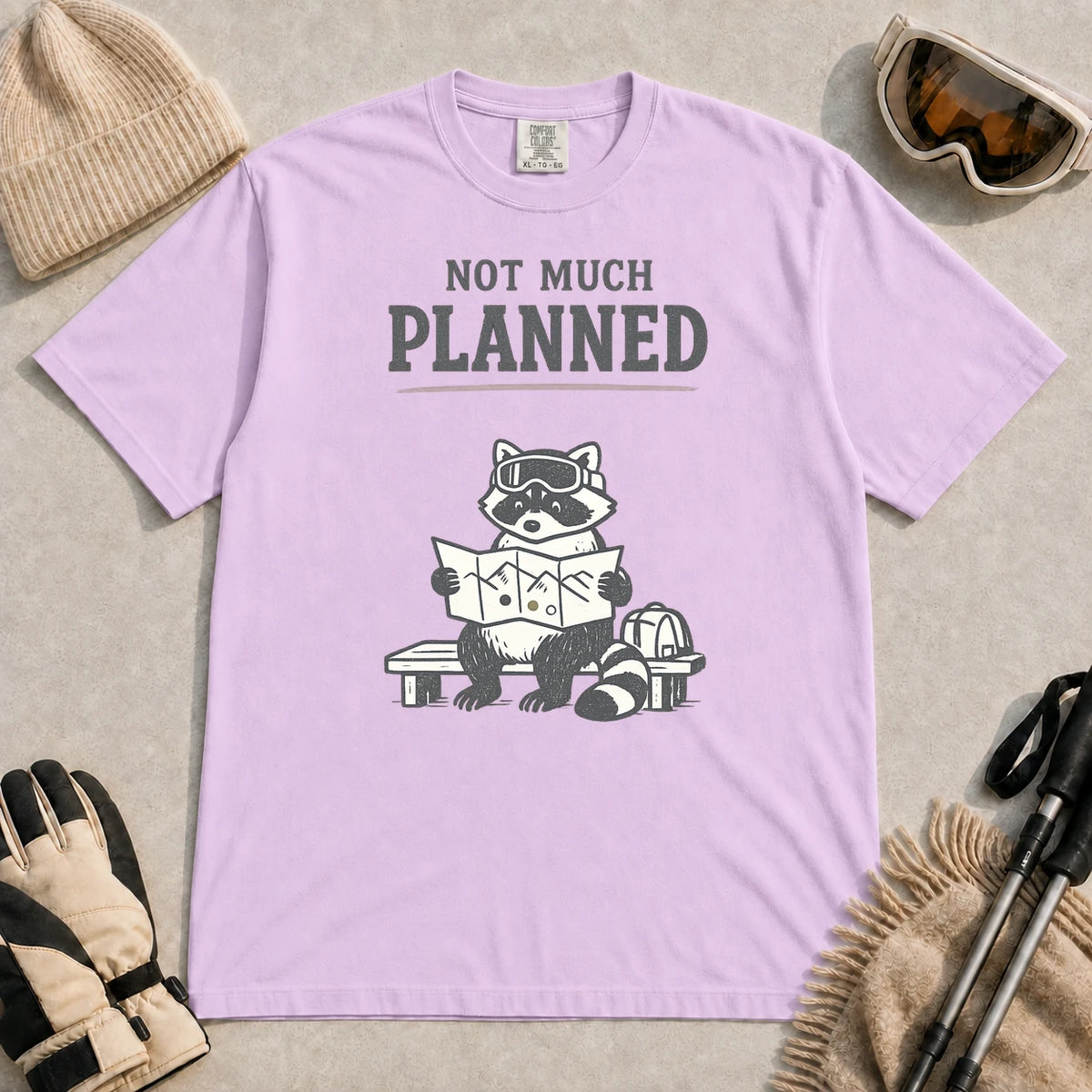 Orchid tee featuring a raccoon in ski goggles sitting on a bench and studying a trail map beside a backpack, with the phrase “Not Much Planned.”