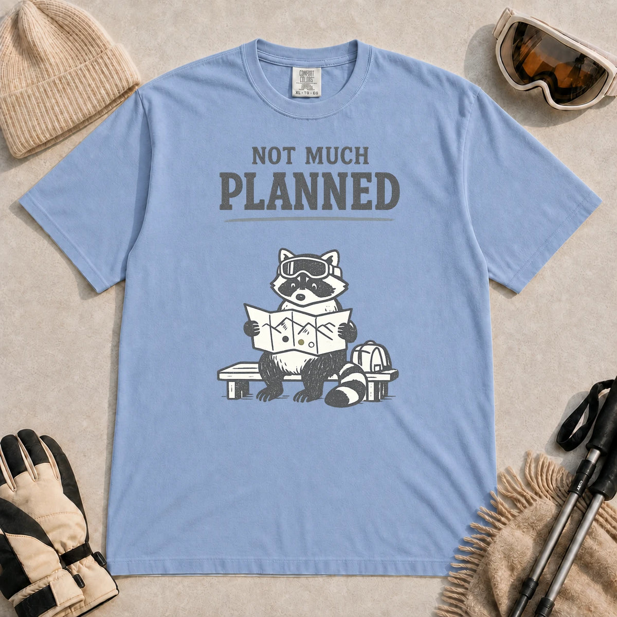 Washed Denim tee featuring a raccoon in ski goggles sitting on a bench and studying a trail map beside a backpack, with the phrase “Not Much Planned.”
