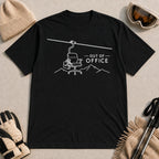 Black tee featuring a minimalist white line-art chairlift carrying an office chair above mountain outlines beside the phrase “Out of Office.”