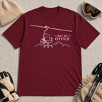 Maroon tee featuring a minimalist white line-art chairlift carrying an office chair above mountain outlines beside the phrase “Out of Office.”