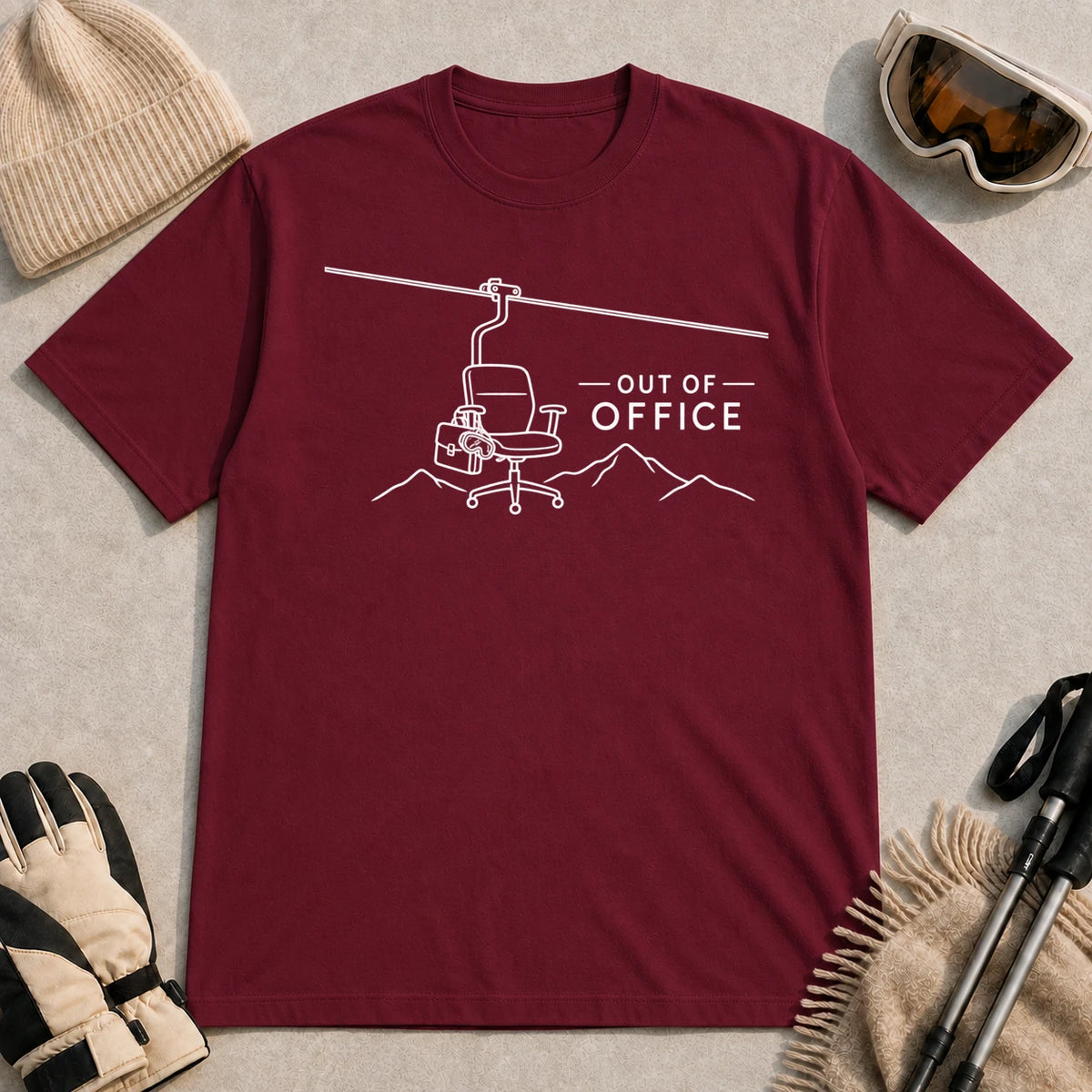 Maroon tee featuring a minimalist white line-art chairlift carrying an office chair above mountain outlines beside the phrase “Out of Office.”