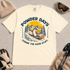 Ivory tee featuring a sunglasses-wearing otter snowboarding through powder beneath a mountain sunset, with the phrases “Powder Days” and “Chasin’ the Good Stuff.”