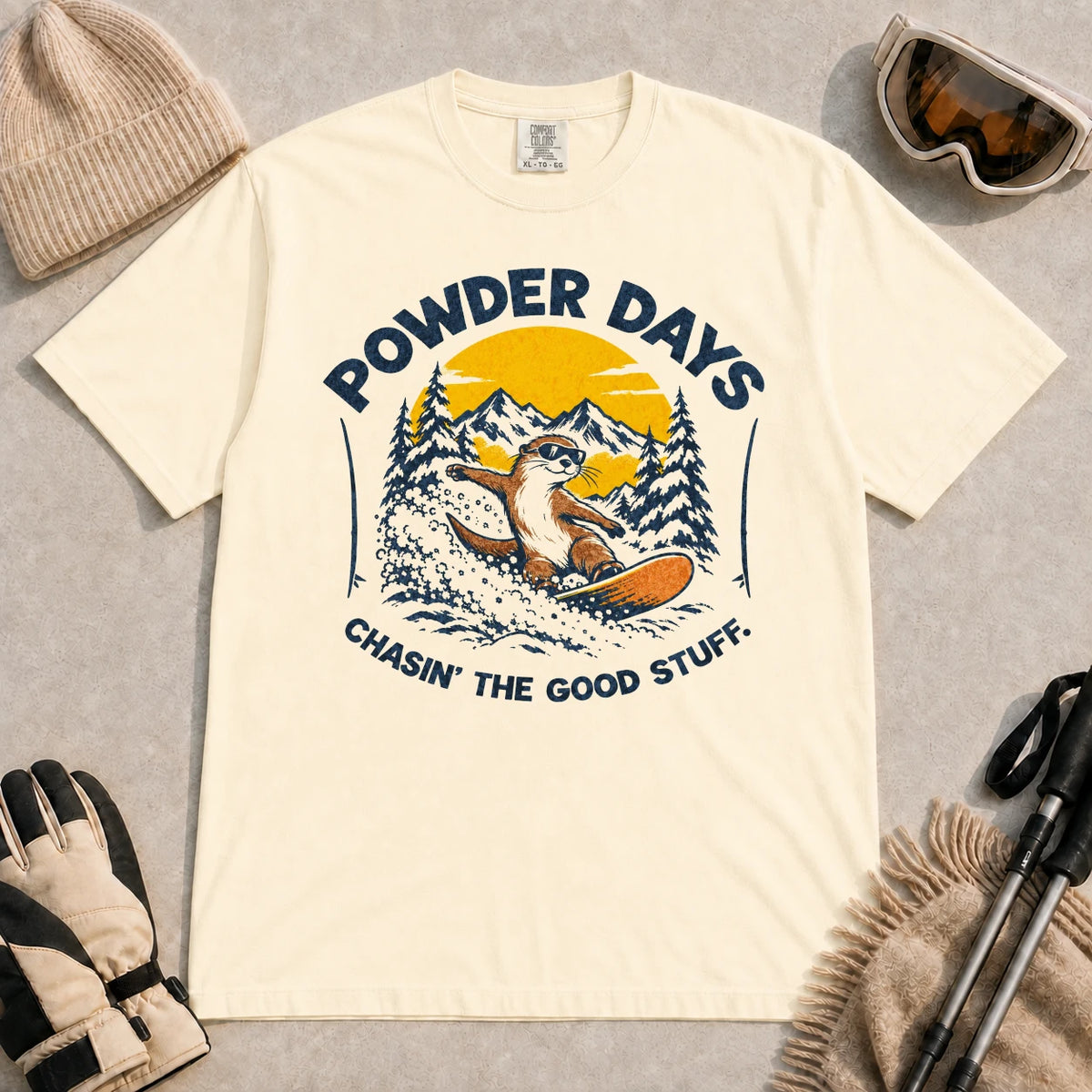 Ivory tee featuring a sunglasses-wearing otter snowboarding through powder beneath a mountain sunset, with the phrases “Powder Days” and “Chasin’ the Good Stuff.”