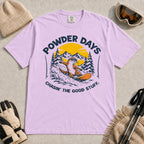 Orchid tee featuring a sunglasses-wearing otter snowboarding through powder beneath a mountain sunset, with the phrases “Powder Days” and “Chasin’ the Good Stuff.”