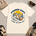 Natural tee featuring a sunglasses-wearing otter snowboarding through powder beneath a mountain sunset, with the phrases “Powder Days” and “Chasin’ the Good Stuff.”