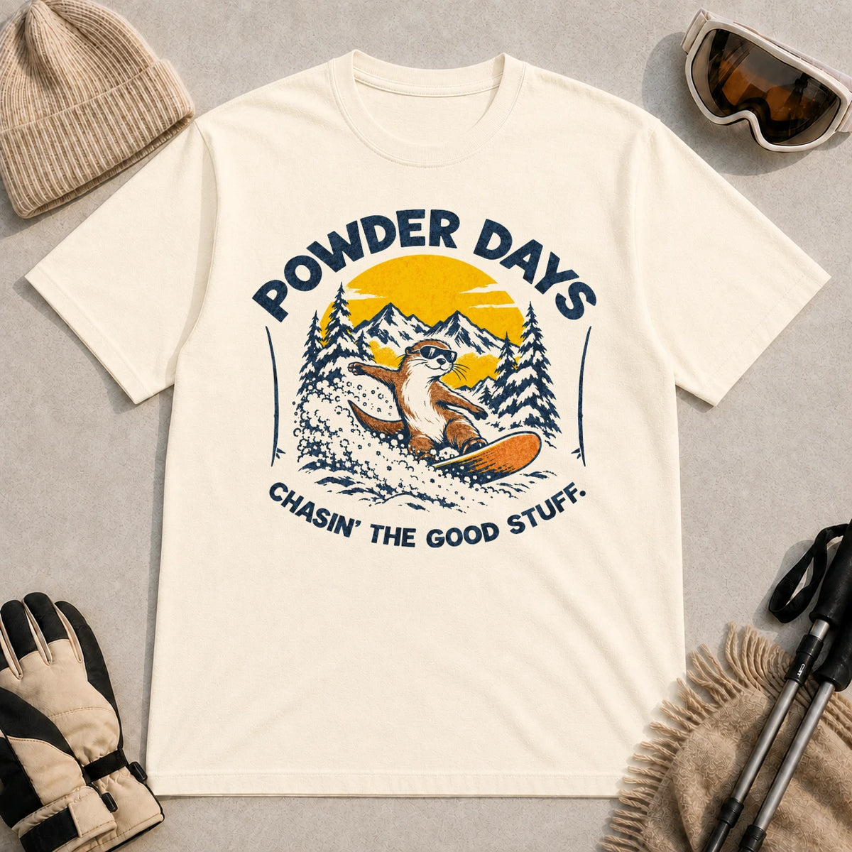 Natural tee featuring a sunglasses-wearing otter snowboarding through powder beneath a mountain sunset, with the phrases “Powder Days” and “Chasin’ the Good Stuff.”