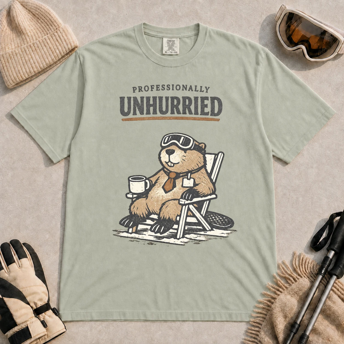 Bay tee featuring a relaxed beaver in ski goggles sitting in a chair with a hot drink, with the phrase “Professionally Unhurried.”
