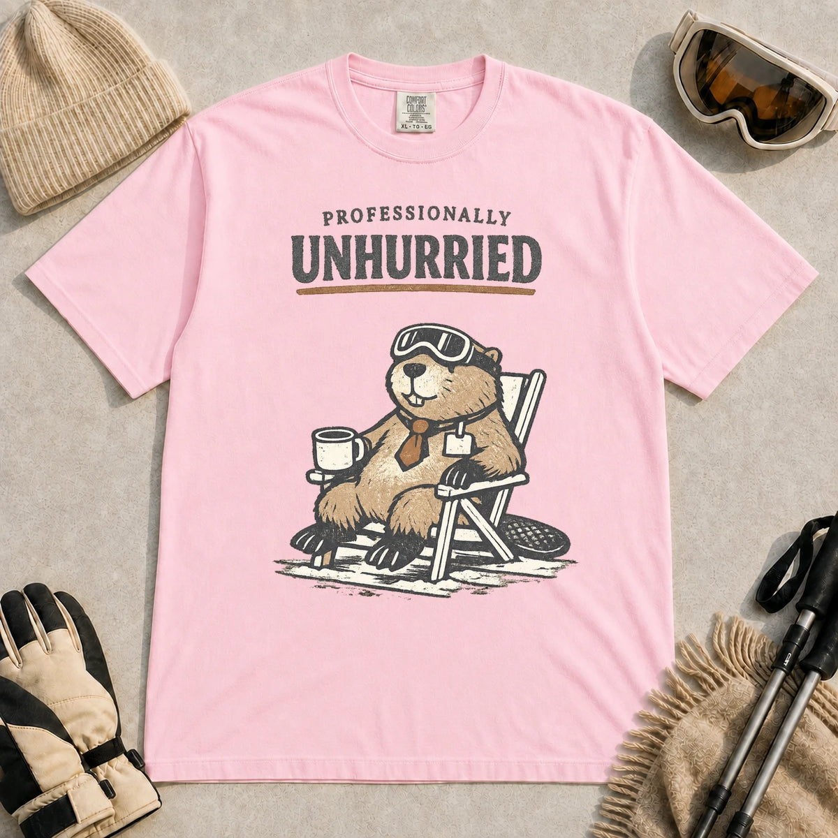 Blossom tee featuring a relaxed beaver in ski goggles sitting in a chair with a hot drink, with the phrase “Professionally Unhurried.”
