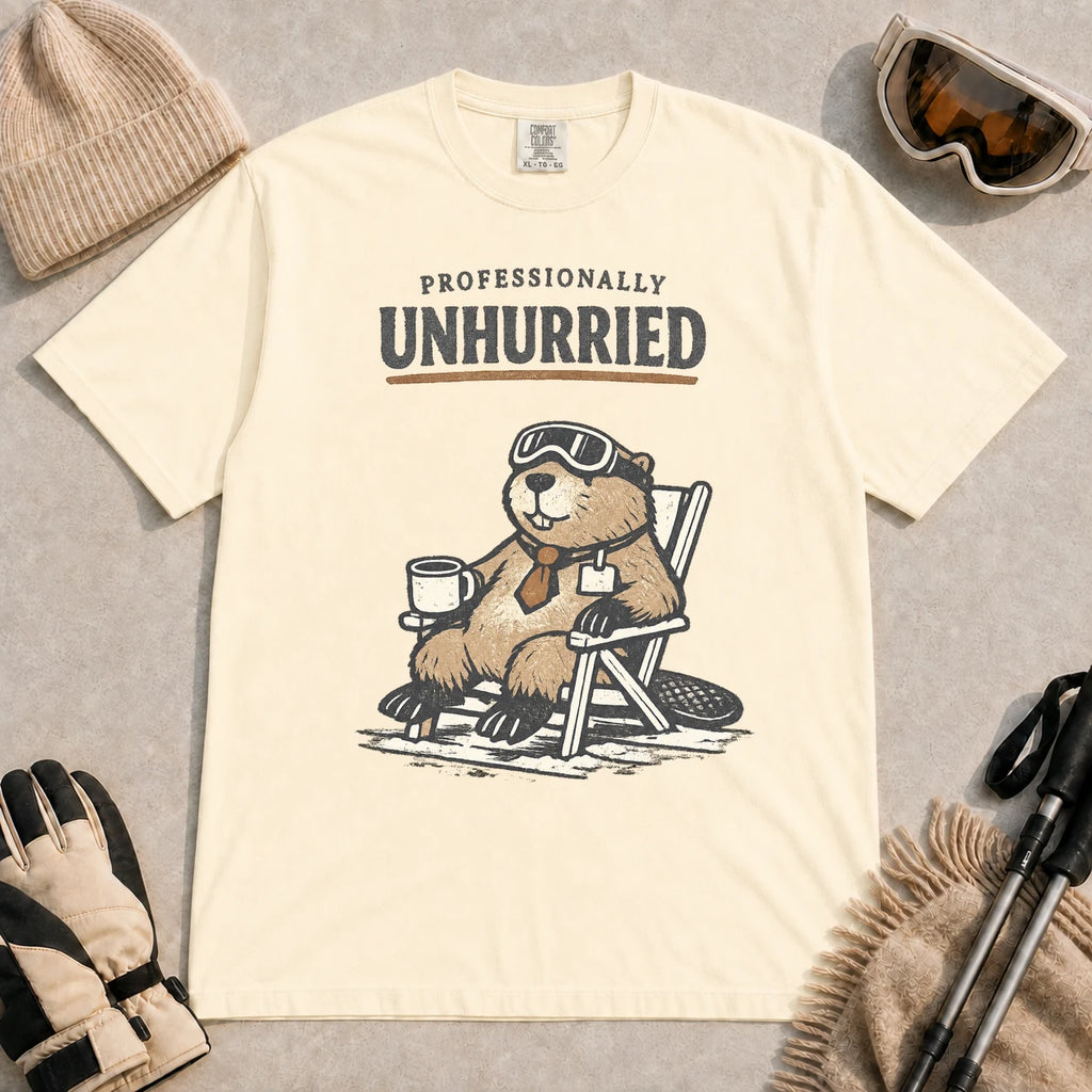 Ivory tee featuring a relaxed beaver in ski goggles sitting in a chair with a hot drink, with the phrase “Professionally Unhurried.”
