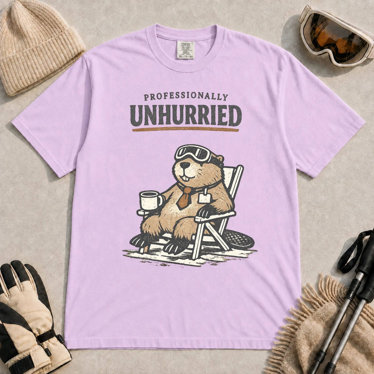 Orchid tee featuring a relaxed beaver in ski goggles sitting in a chair with a hot drink, with the phrase “Professionally Unhurried.”