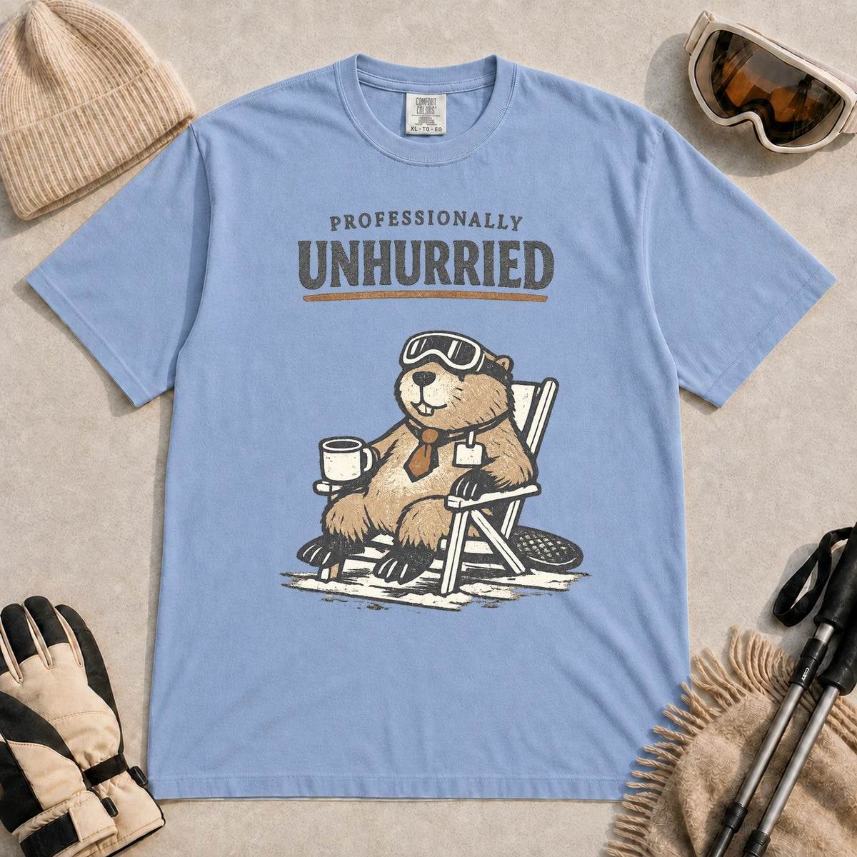 Washed Denim tee featuring a relaxed beaver in ski goggles sitting in a chair with a hot drink, with the phrase “Professionally Unhurried.”