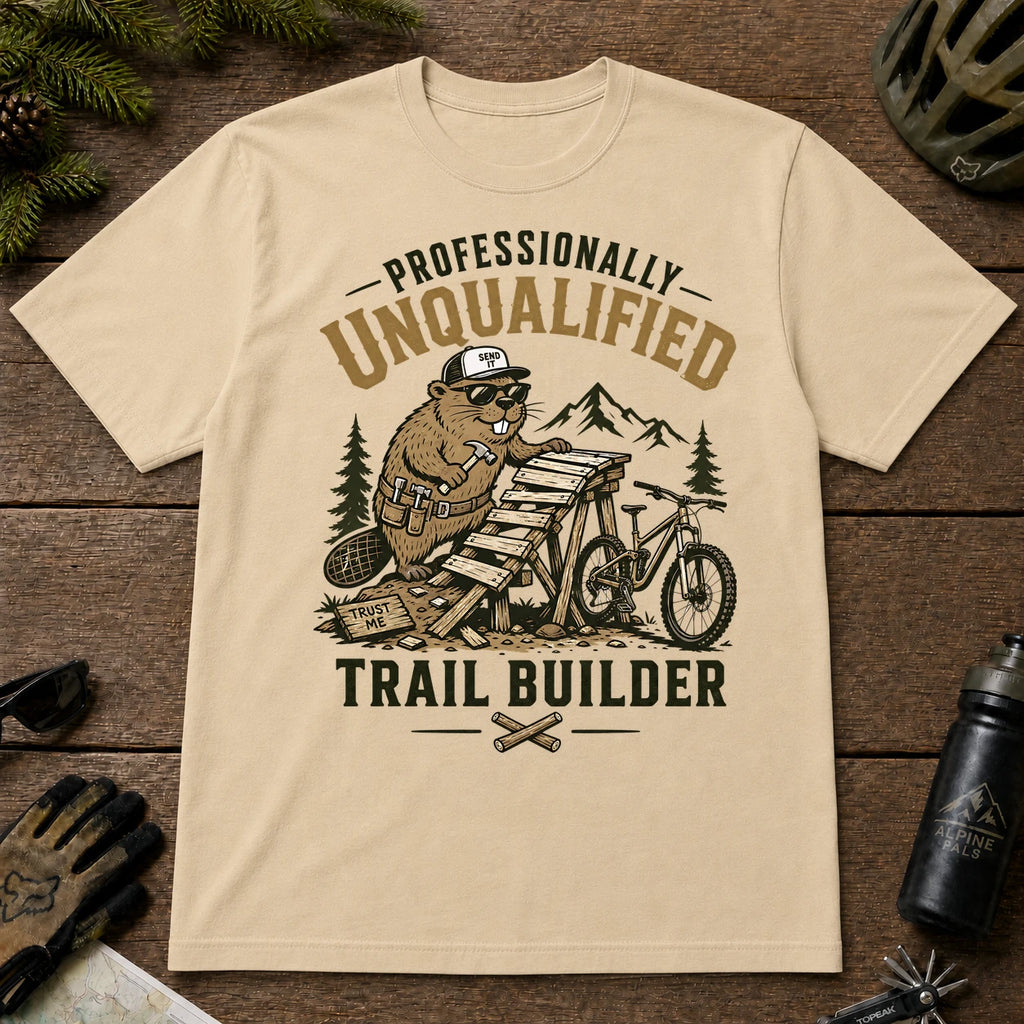 Sand tee featuring a sunglasses-wearing beaver building a wooden mountain bike ramp beside a bike, with the phrase “Professionally Unqualified Trail Builder.”