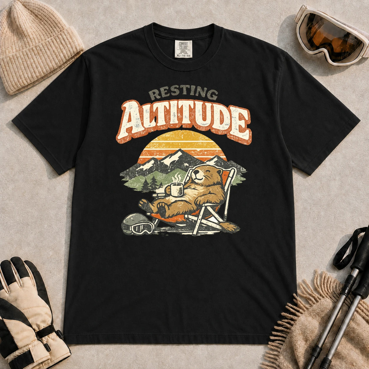 Black tee featuring a relaxed marmot lounging in a chair with a hot drink beside ski goggles, backed by mountains and a retro sunset with the phrase “Resting Altitude.”