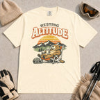 Ivory tee featuring a relaxed marmot lounging in a chair with a hot drink beside ski goggles, backed by mountains and a retro sunset with the phrase “Resting Altitude.”