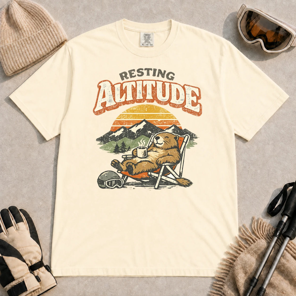 Ivory tee featuring a relaxed marmot lounging in a chair with a hot drink beside ski goggles, backed by mountains and a retro sunset with the phrase “Resting Altitude.”