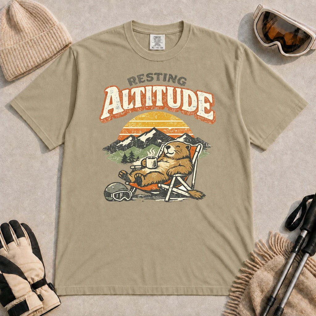 Khaki tee featuring a relaxed marmot lounging in a chair with a hot drink beside ski goggles, backed by mountains and a retro sunset with the phrase “Resting Altitude.”