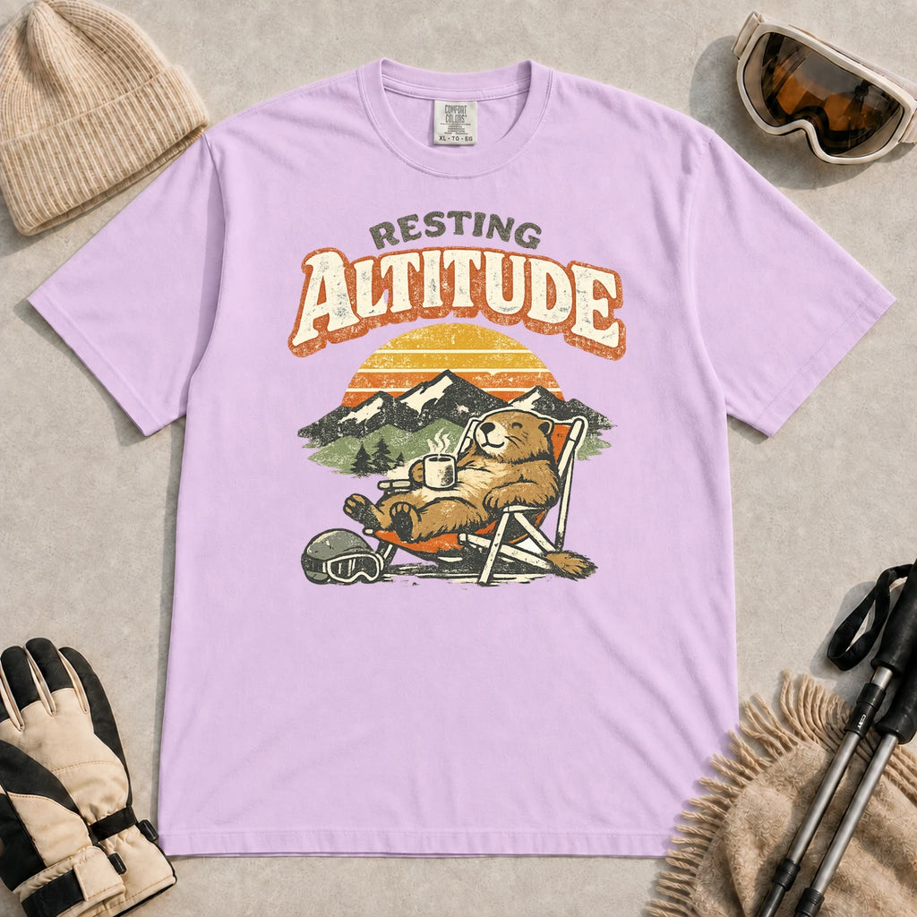 Orchid tee featuring a relaxed marmot lounging in a chair with a hot drink beside ski goggles, backed by mountains and a retro sunset with the phrase “Resting Altitude.”