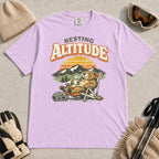 Orchid tee featuring a relaxed marmot lounging in a chair with a hot drink beside ski goggles, backed by mountains and a retro sunset with the phrase “Resting Altitude.”