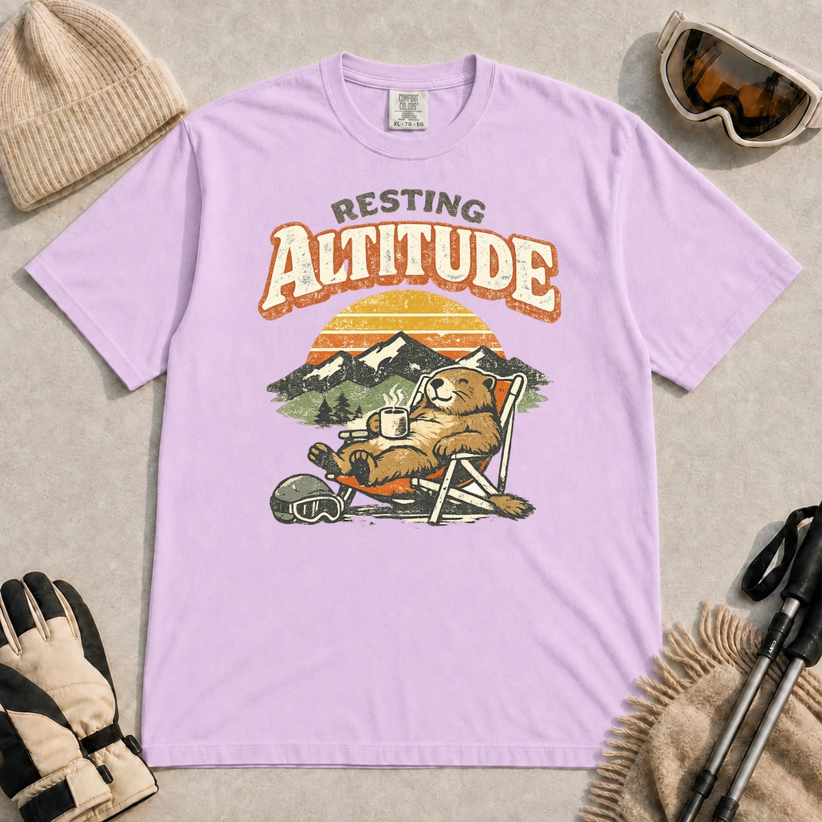 Orchid tee featuring a relaxed marmot lounging in a chair with a hot drink beside ski goggles, backed by mountains and a retro sunset with the phrase “Resting Altitude.”