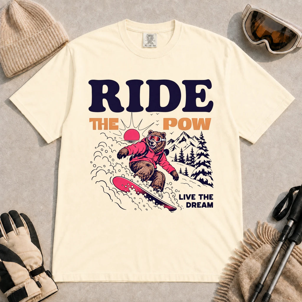 Ivory tee featuring a bear snowboarding through powder in a retro mountain scene with the phrases “Ride the Pow” and “Live the Dream.”