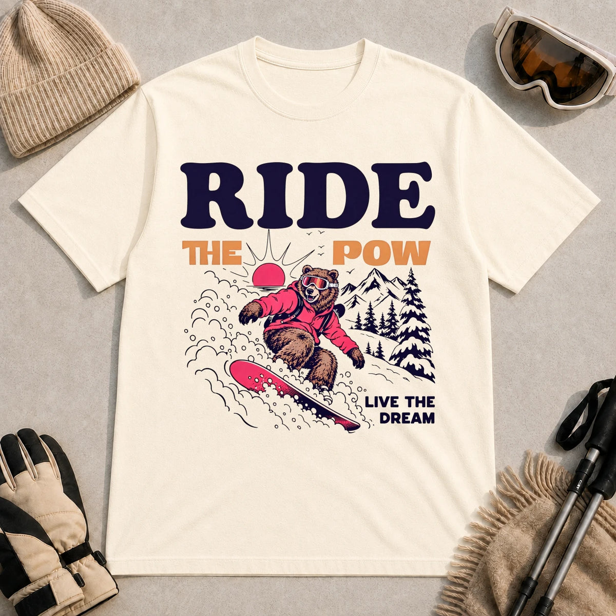 Natural tee featuring a bear snowboarding through powder in a retro mountain scene with the phrases “Ride the Pow” and “Live the Dream.”