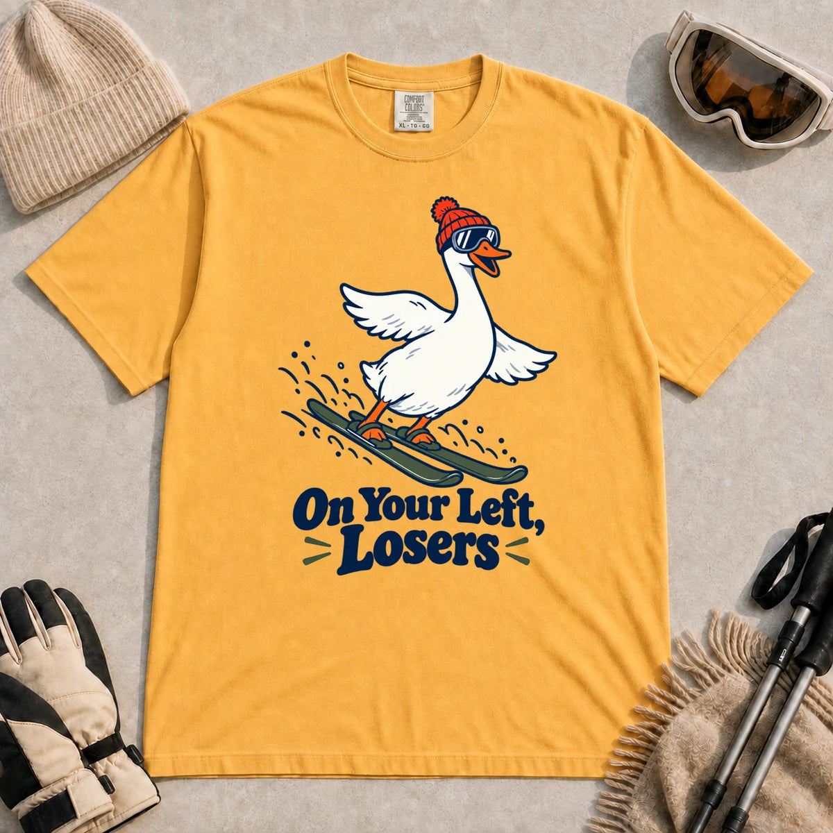 Mustard tee featuring a playful goose skiing in goggles and a beanie above the phrase “On Your Left, Losers.”