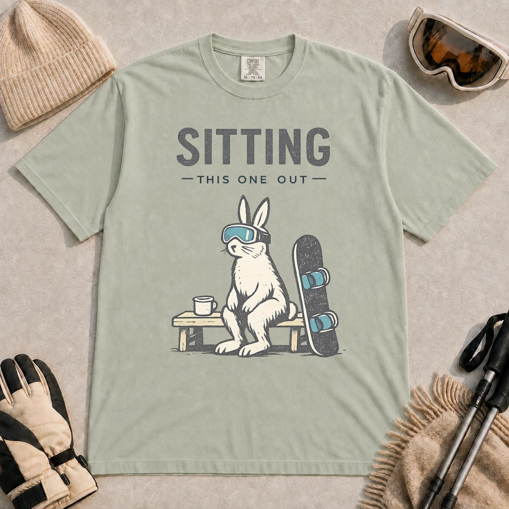 Bay tee featuring a bunny in ski goggles sitting on a bench beside a snowboard and hot drink with the phrase “Sitting This One Out.”