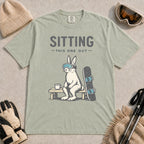 Bay tee featuring a bunny in ski goggles sitting on a bench beside a snowboard and hot drink with the phrase “Sitting This One Out.”