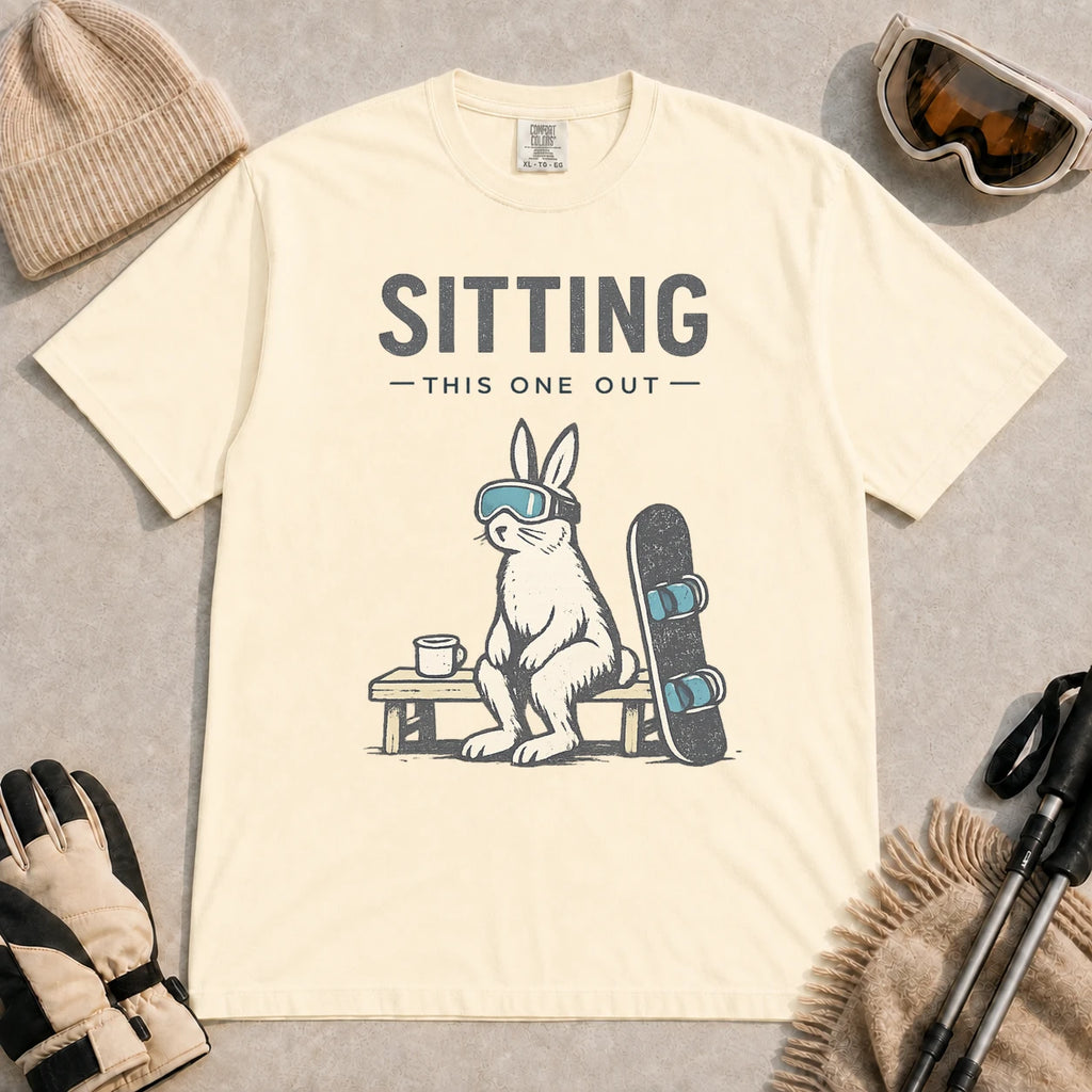 Ivory tee featuring a bunny in ski goggles sitting on a bench beside a snowboard and hot drink with the phrase “Sitting This One Out.”