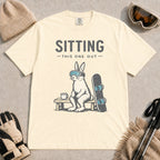 Ivory tee featuring a bunny in ski goggles sitting on a bench beside a snowboard and hot drink with the phrase “Sitting This One Out.”